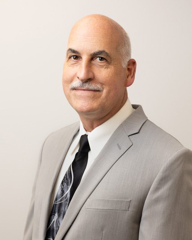 A professional P.E. headshot of a bald man with a mustache, wearing a grey suit and black tie.