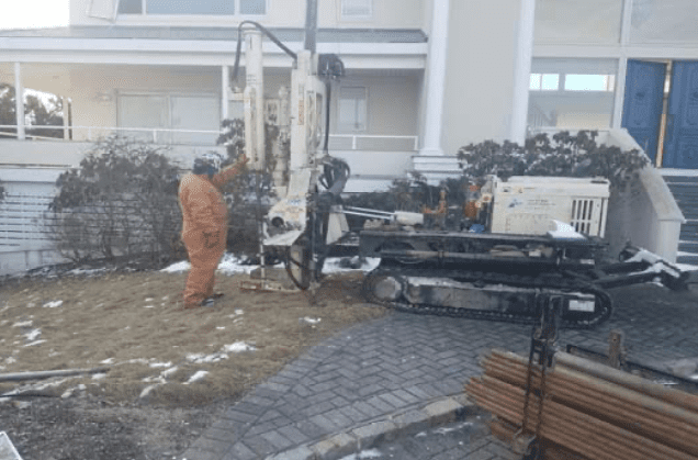 A worker in protective gear operating a drilling rig near a residential building as part of an Environmental Site Assessment phase I & II due diligence process.