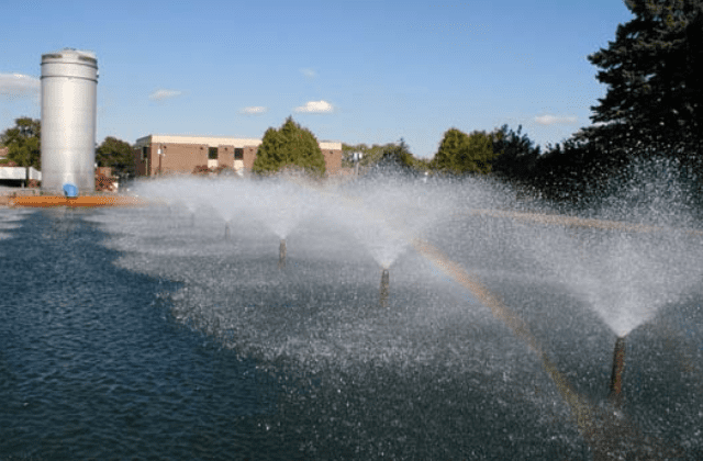 Spray fountains operating in a pond with a silo and buildings in the background, incorporating elements of water system design.