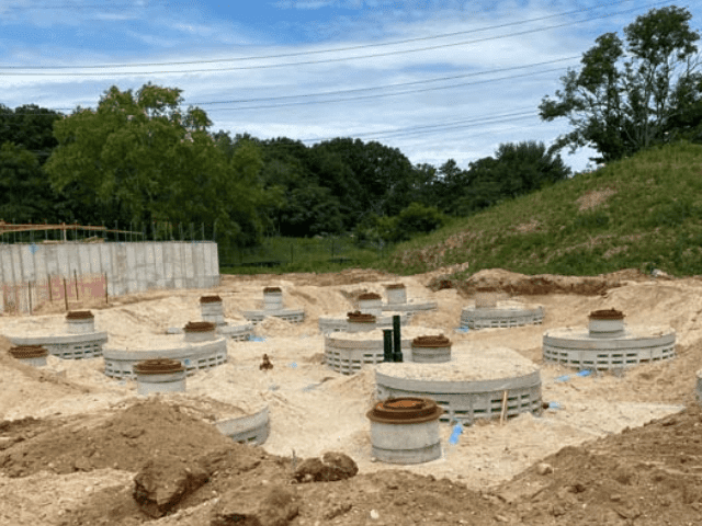 Construction site with concrete foundations and cylindrical structures, likely for road and drainage infrastructure development, surrounded by dirt and sand.