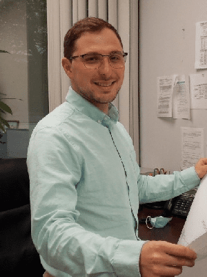 Man with glasses, Jim Ferraiuolo, smiling at a desk in an office environment.