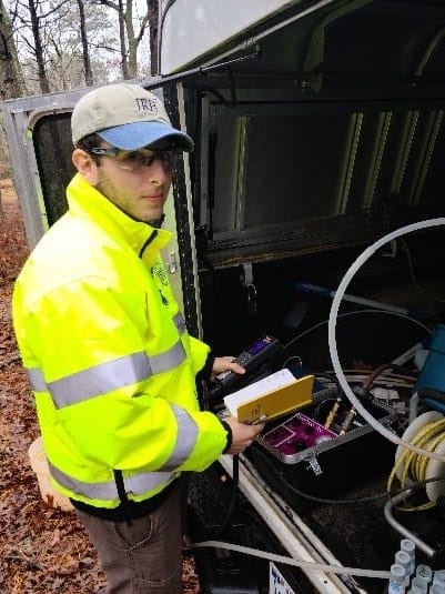 A technician, also an Environmental Scientist named Jim Ferraiuolo, wearing a high-visibility jacket and cap operating equipment inside a utility vehicle.