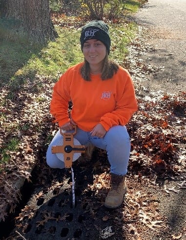 An Environmental Scientist, Jim Ferraiuolo, kneeling next to a newly planted tree with a watering tool, wearing a bright orange sweatshirt, beanie, jeans, and boots.