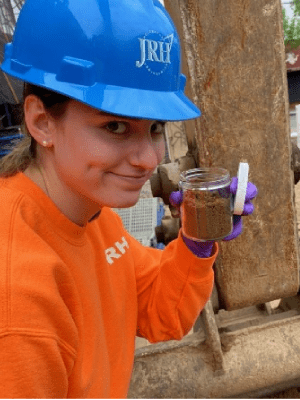 Environmental Scientist in a hard hat holding a jar of soil sample at a construction or drilling site.