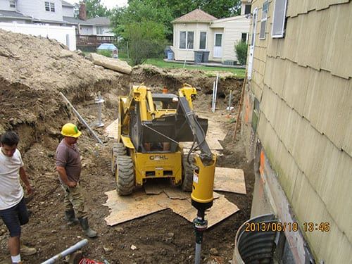 Construction workers operating a JRH skid-steer loader near an excavation site by a residential building.